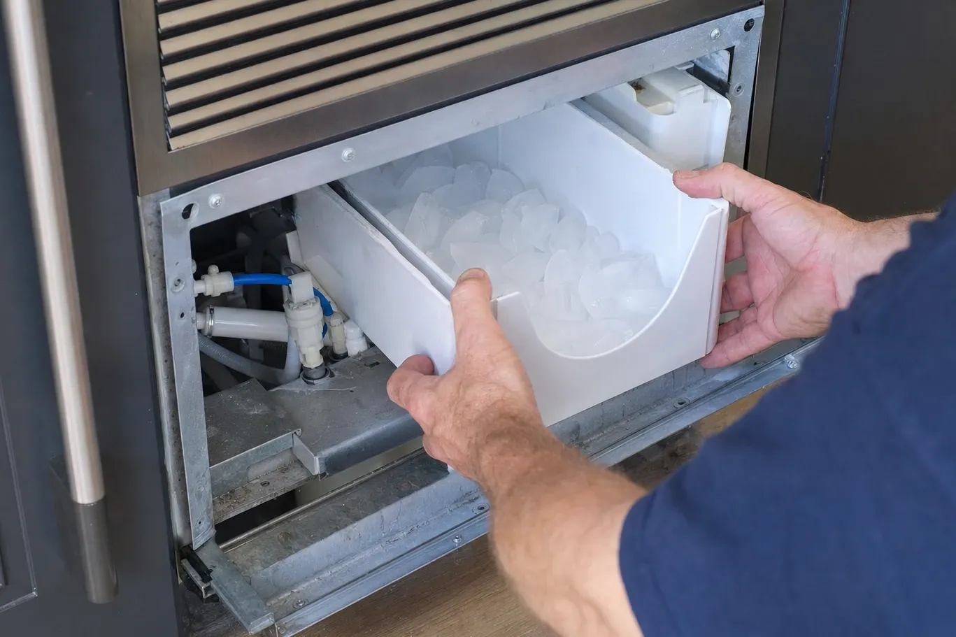 Hands drawing a storage bin heaped with ice cubes out of an undercounter ice machine, an inline water filter and blue supply tubing visible in the bay behind it